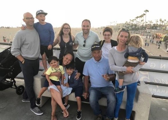 A large family group gathered by the beach pier with palm trees behind them